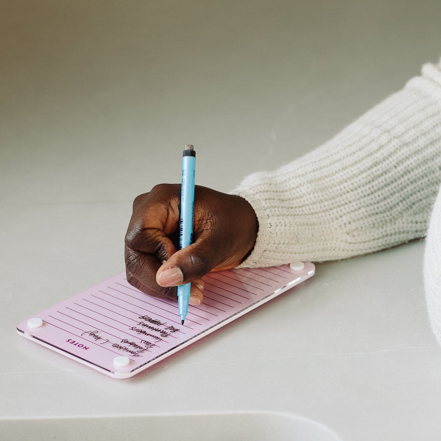 Someone wearing a sweater writes meeting notes on Monkey and Pebbles' Lined Notes 59 pink notepad with a blue pen, resting their hand on a light-colored surface.