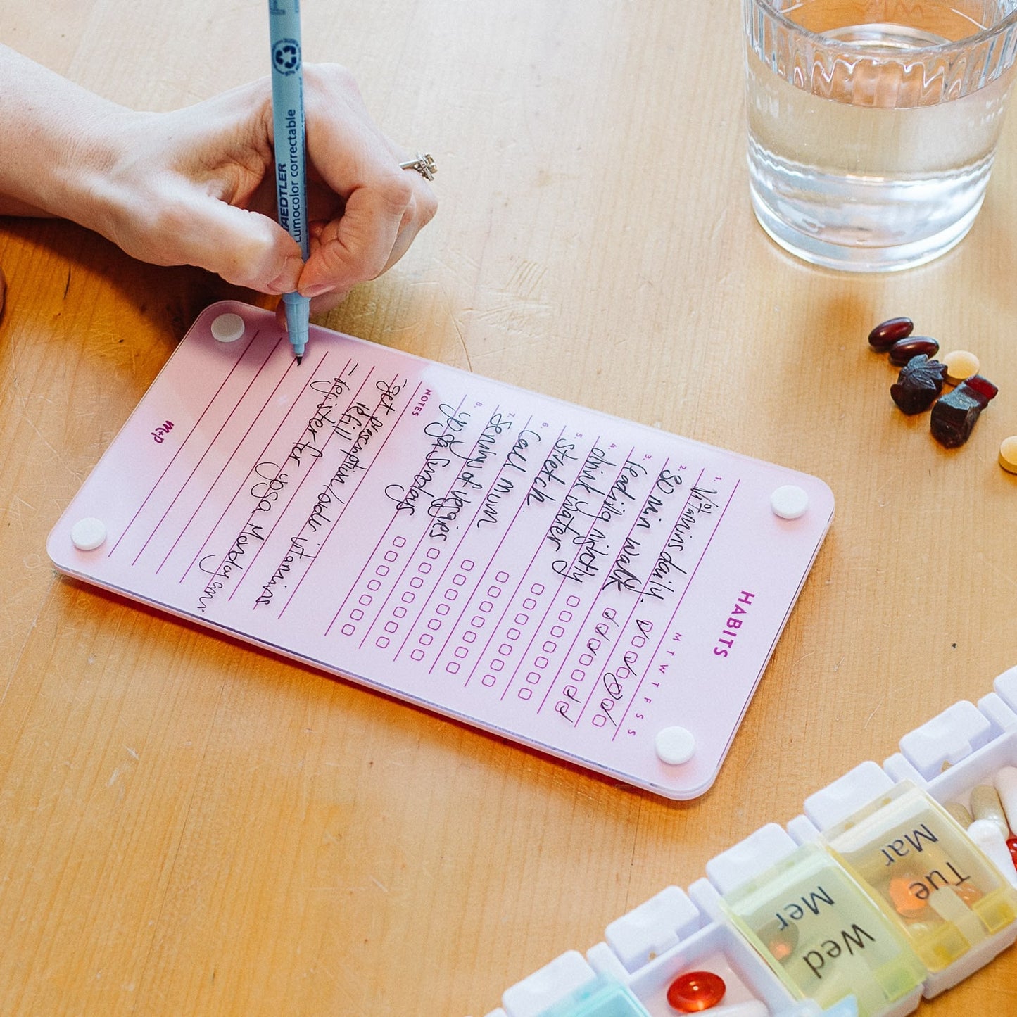 Someone uses a blue pen to fill out the Monkey and Pebbles Weekly Habits 59 pink tracker at a wooden table, surrounded by assorted pills, a pill organizer, and tools for setting goals and planning routines.