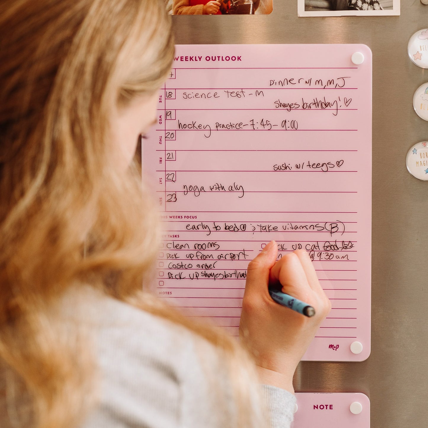 A person writes with a pencil on the pink Weekly Outlook 811 planner by Monkey and Pebbles, attached to the fridge, organizing events like a science test, dinner, and hockey practice with daily notes to boost productivity.
