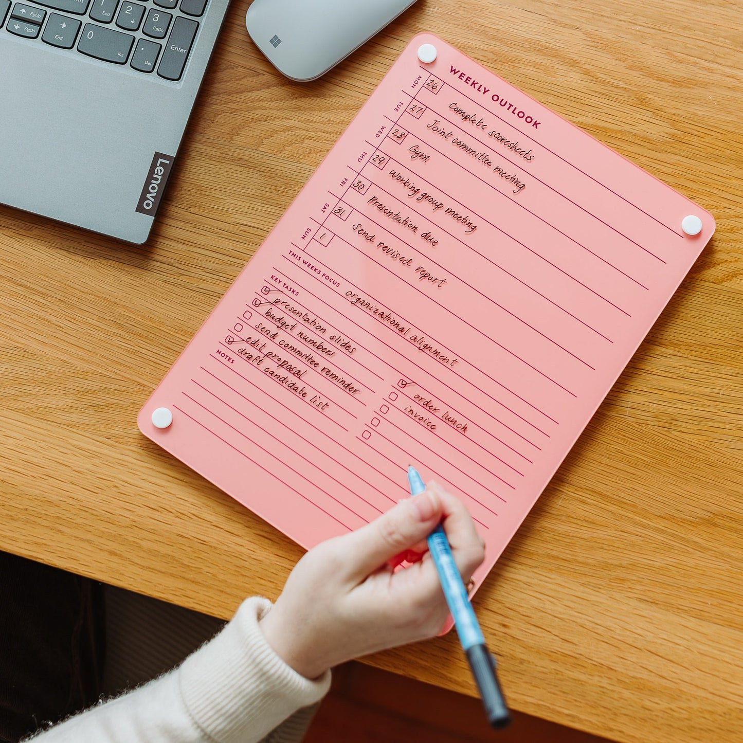 At a wooden desk beside a Lenovo laptop, someone writes on the Monkey and Pebbles Weekly Outlook 811 pink planner with a blue pen, organizing their weekly tasks and reminders to boost productivity.