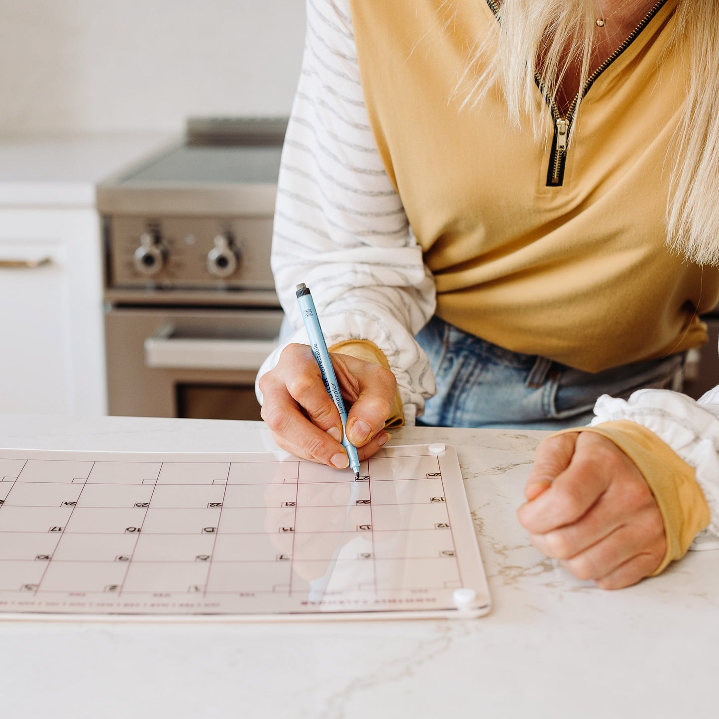 A person writes on the Monkey and Pebbles Monthly Calendar 1411 placed on the kitchen counter, with a stove in the background, while wearing a yellow and white long-sleeve shirt.