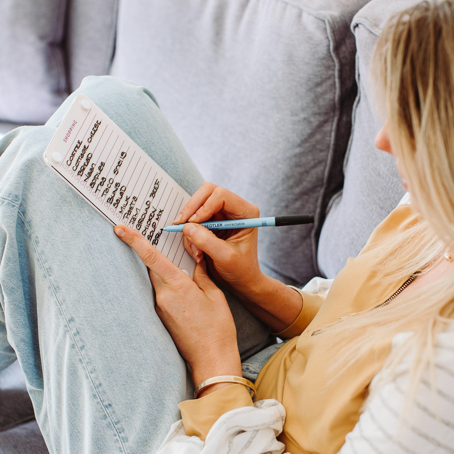 A person writes a meal plan on the Monkey and Pebbles Shopping List 59 notepad, jotting down essentials like coffee, granola, cereal, and bread while seated on a couch. Only their hands and part of their face are in view.