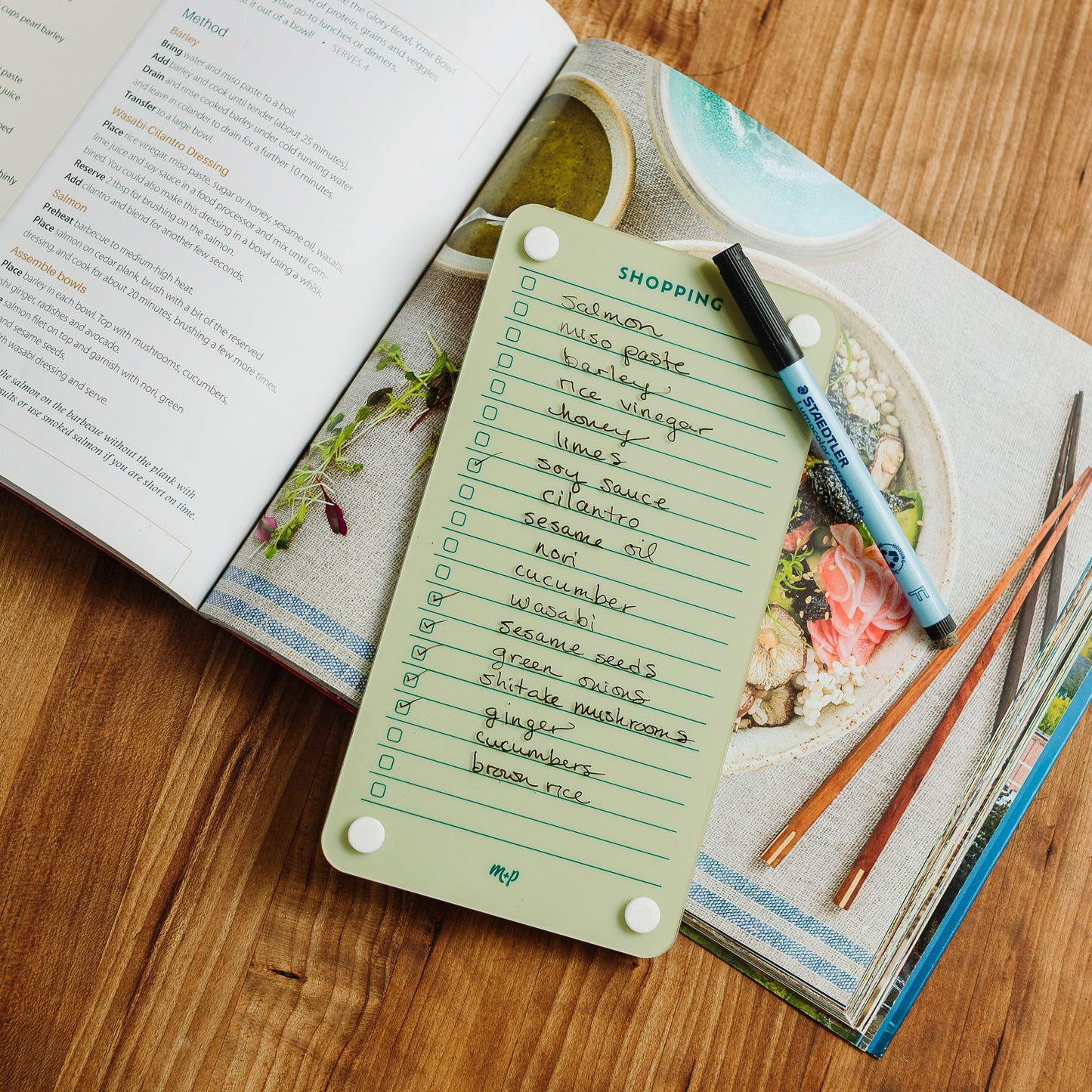 The Monkey and Pebbles Shopping List 59, a green grocery list board with marker, rests on an open cookbook displaying an Asian recipe. Items listed include soy sauce, miso paste, sesame oil, and ginger on a wooden table.
