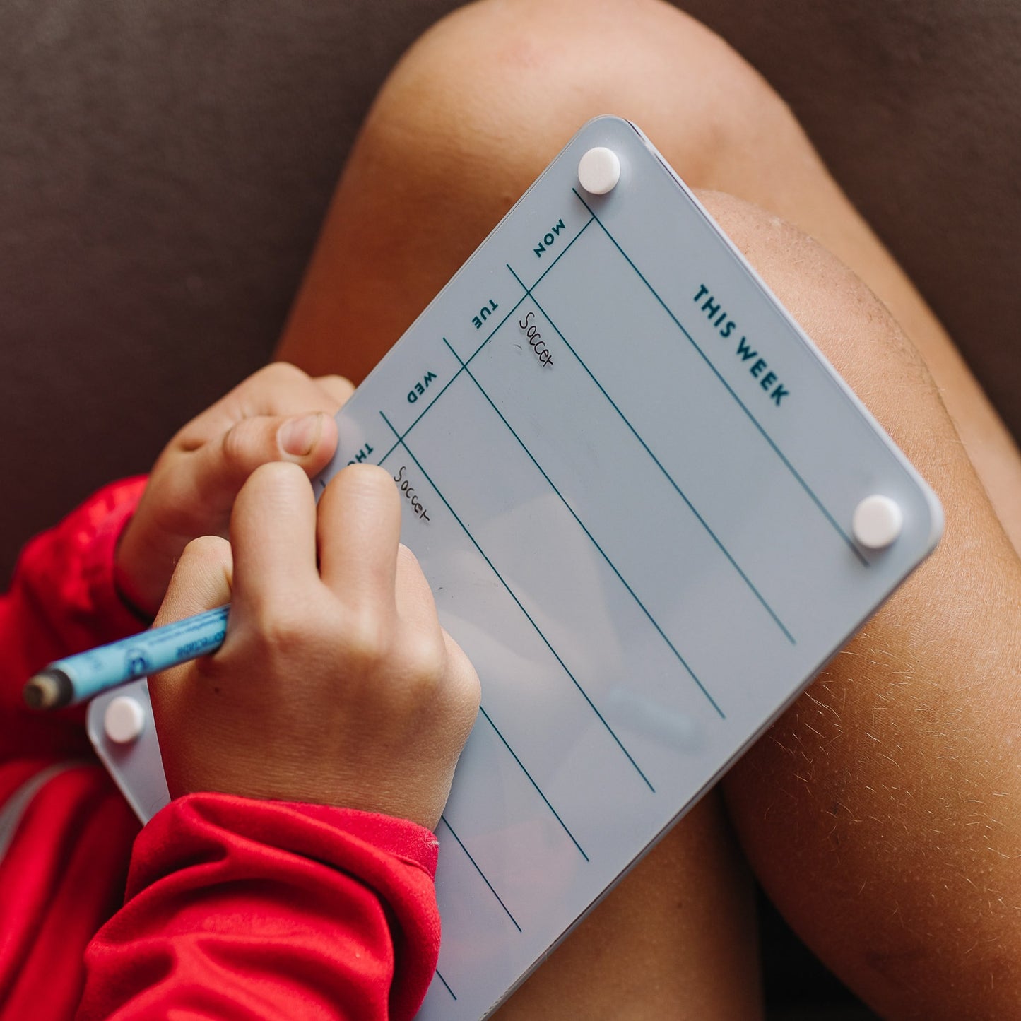 A person holds the Monkey and Pebbles "This Week 69" blue weekly organizer on their knee, writing with a pencil. The planner has columns for each day and is blank except for notes on Monday and Sunday.