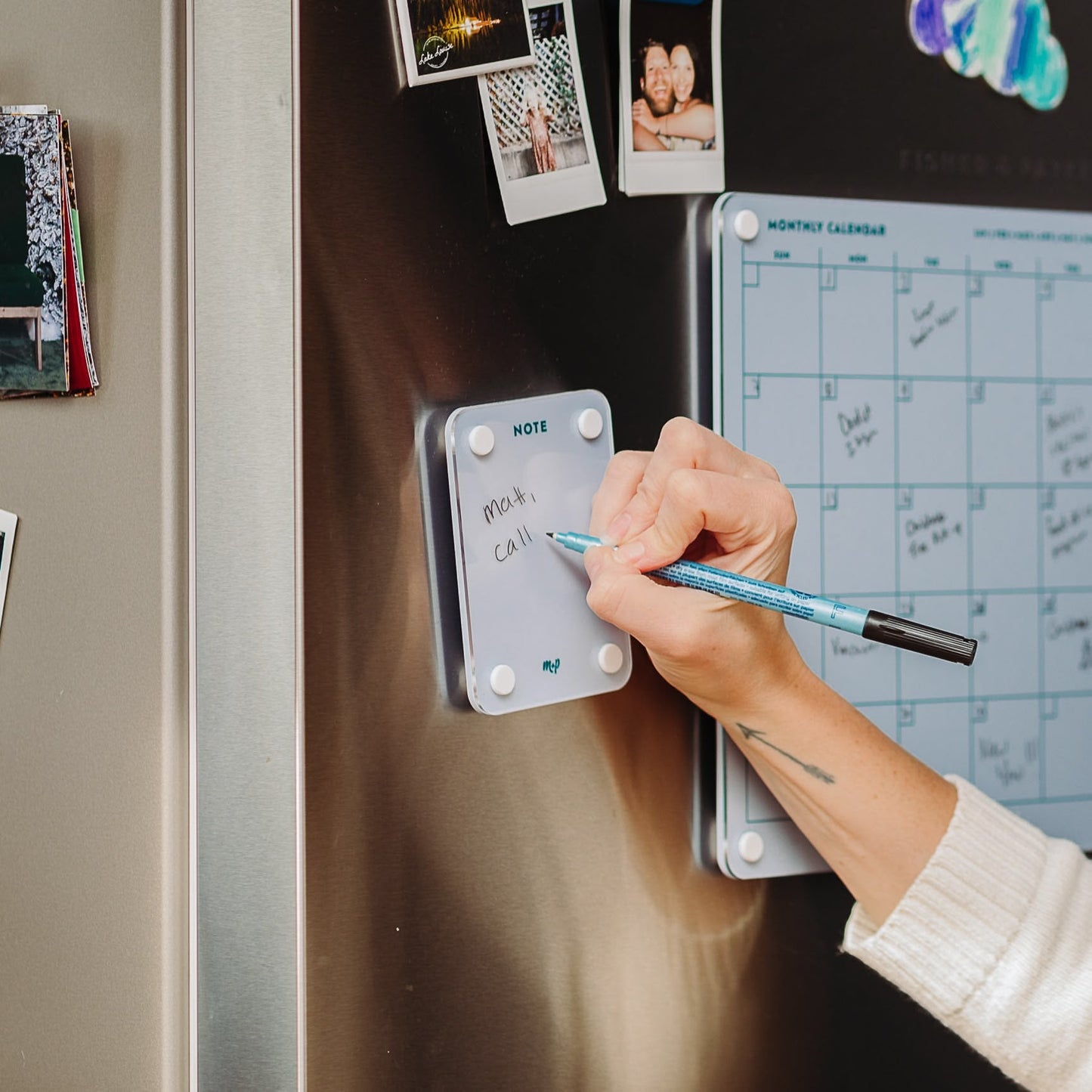Someone writes "meals" and "call" on a Monkey and Pebbles Little Note 35 sticky note attached to the fridge, next to a blue monthly calendar and several photos.