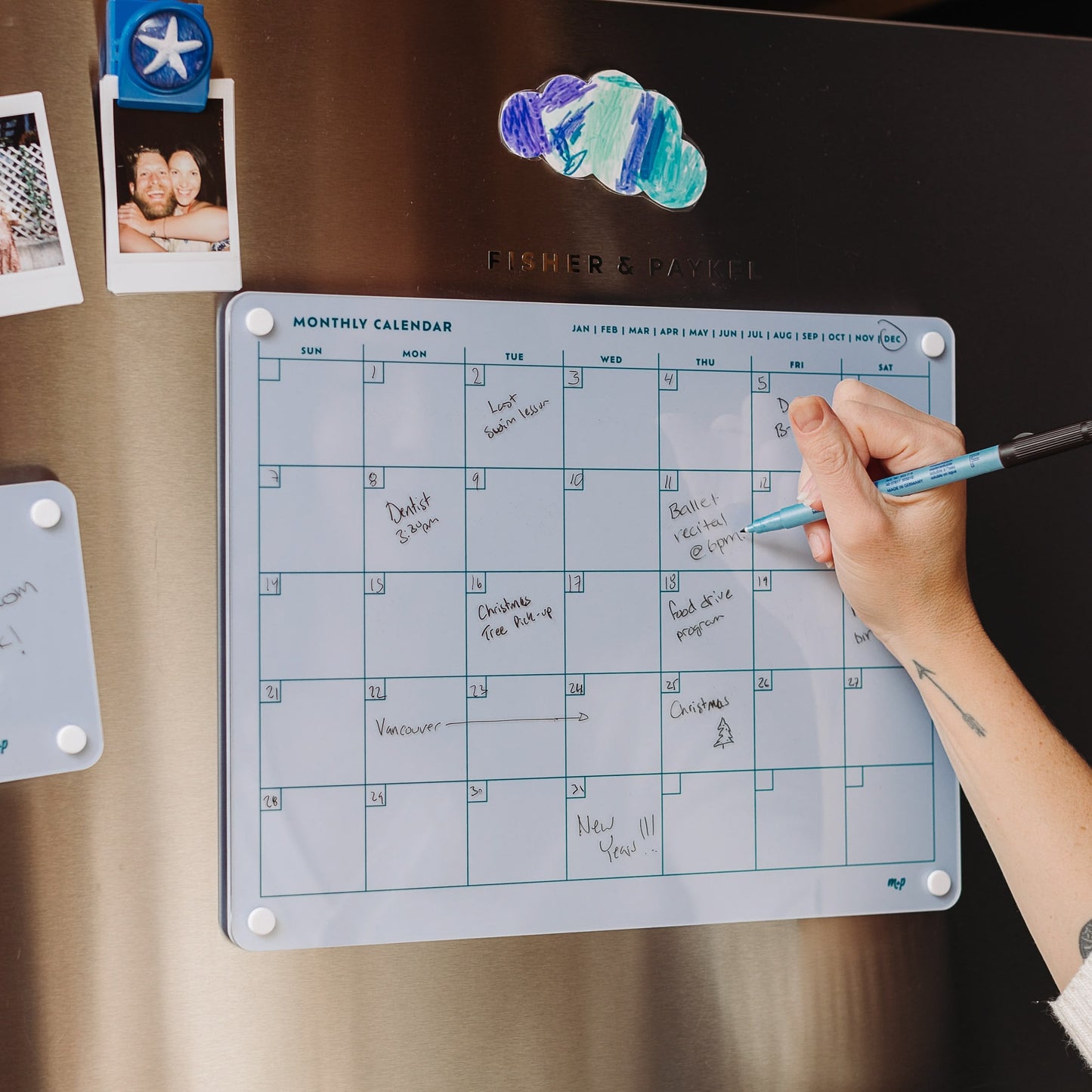 A person writes on the Monkey and Pebbles Monthly Calendar 1411, a blue dry-erase planner attached to a stainless steel fridge, surrounded by family photos and featuring handwritten notes for organizing events.