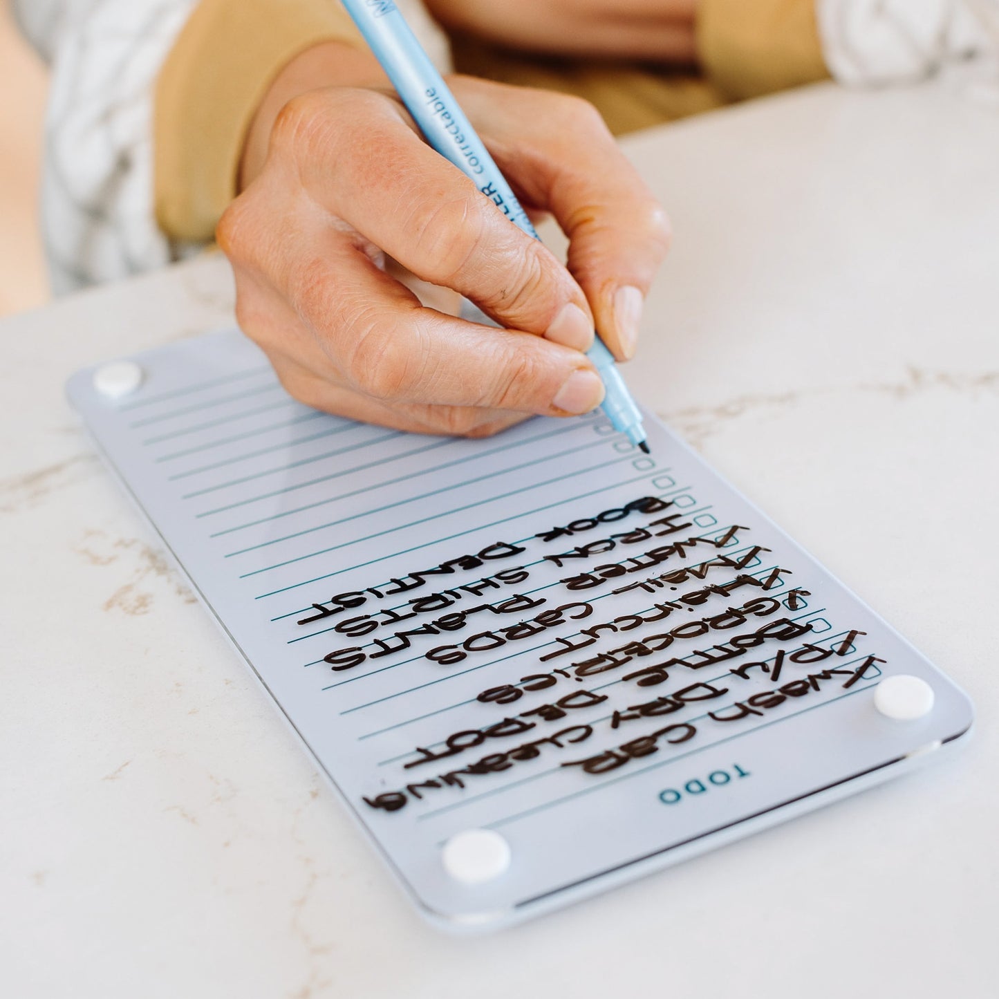 A hand holding a blue pen checks off tasks on the Monkey and Pebbles Todo List 59, a reusable organizer with lined sections and "TODO" at the bottom, resting on a smooth surface.