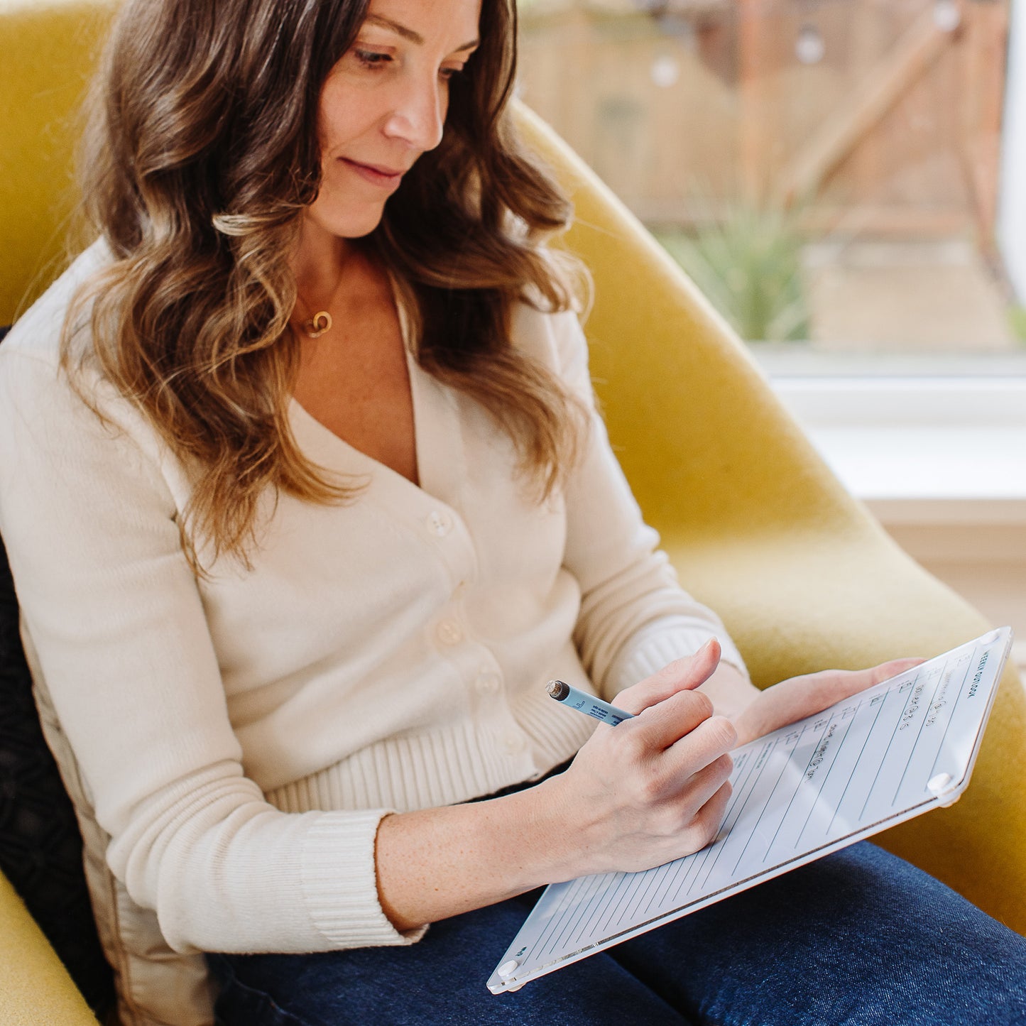 Seated on a yellow chair in a cream sweater and jeans, a woman with wavy brown hair stays focused as she fills out her Weekly Outlook 811 planner, organizing her tasks for the week to enhance productivity.
