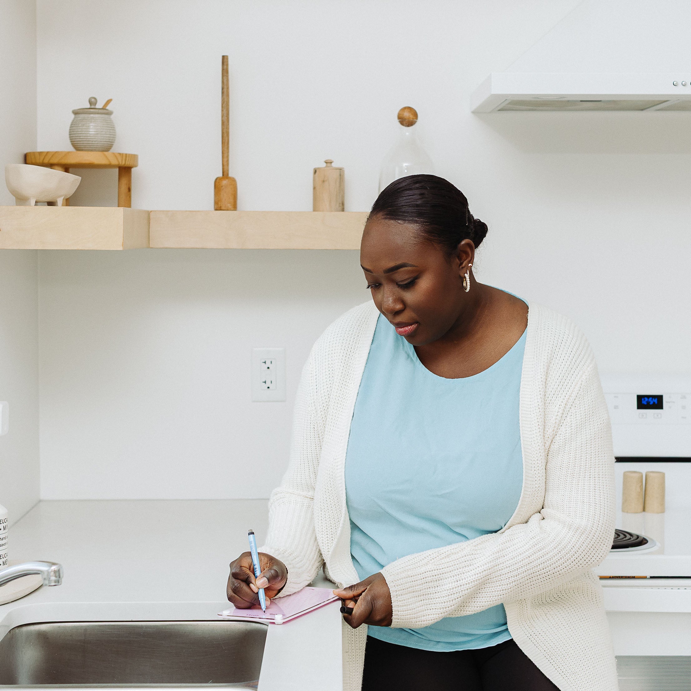 In a modern kitchen, a woman in a light blue shirt and cardigan leans against the counter, jotting meeting notes and doodles in her Monkey and Pebbles Lined Notes 59 notebook. Shelves with jars and dishes are visible in the background.