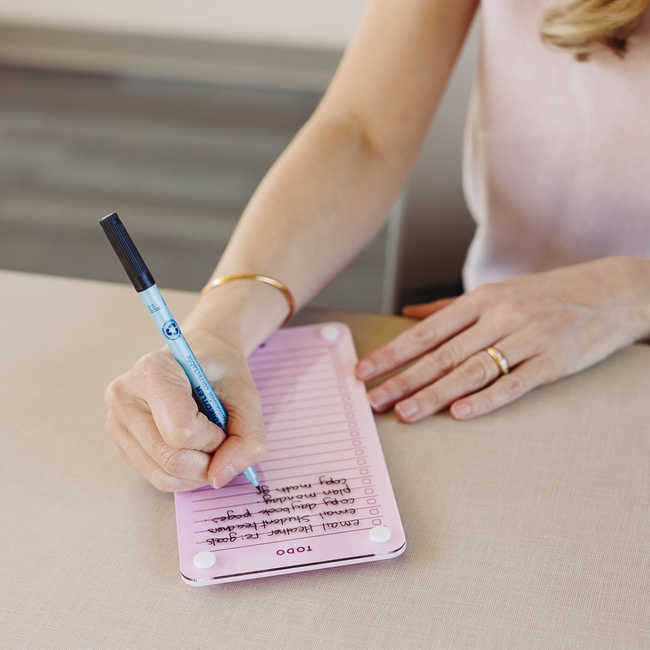 A person wearing a gold ring and bracelet writes a to-do list on the Monkey and Pebbles Todo List 59 pink notepad with a blue pen while sitting at a beige table, using it as their daily planner.