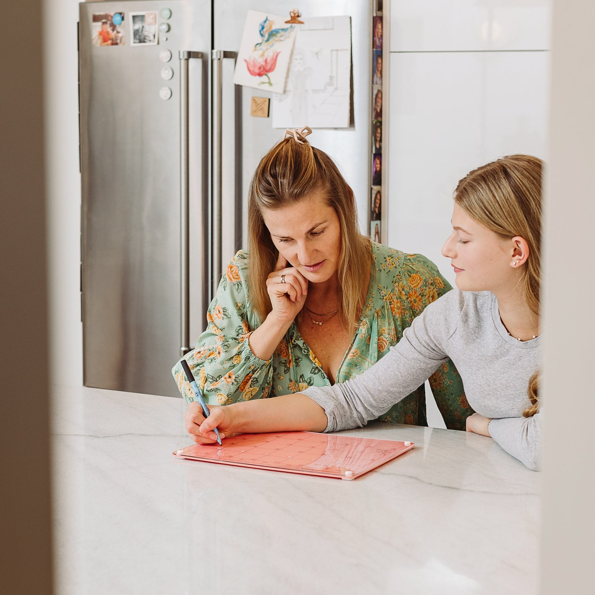 A woman in a green floral blouse and a teenage girl in a gray shirt sit at a kitchen counter, using the Monkey and Pebbles Monthly Calendar 1411 tablet to organize plans, with a photo-covered fridge visible behind them.