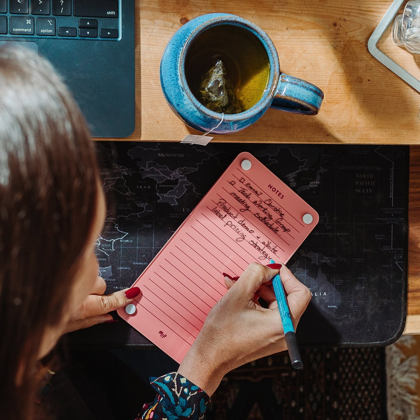 A person writes meeting notes on the Monkey and Pebbles Lined Notes 59 pink notepad at a desk with a laptop, blue mug of tea, and black map mouse pad. The top-down photo shows their hand holding a turquoise pen.
