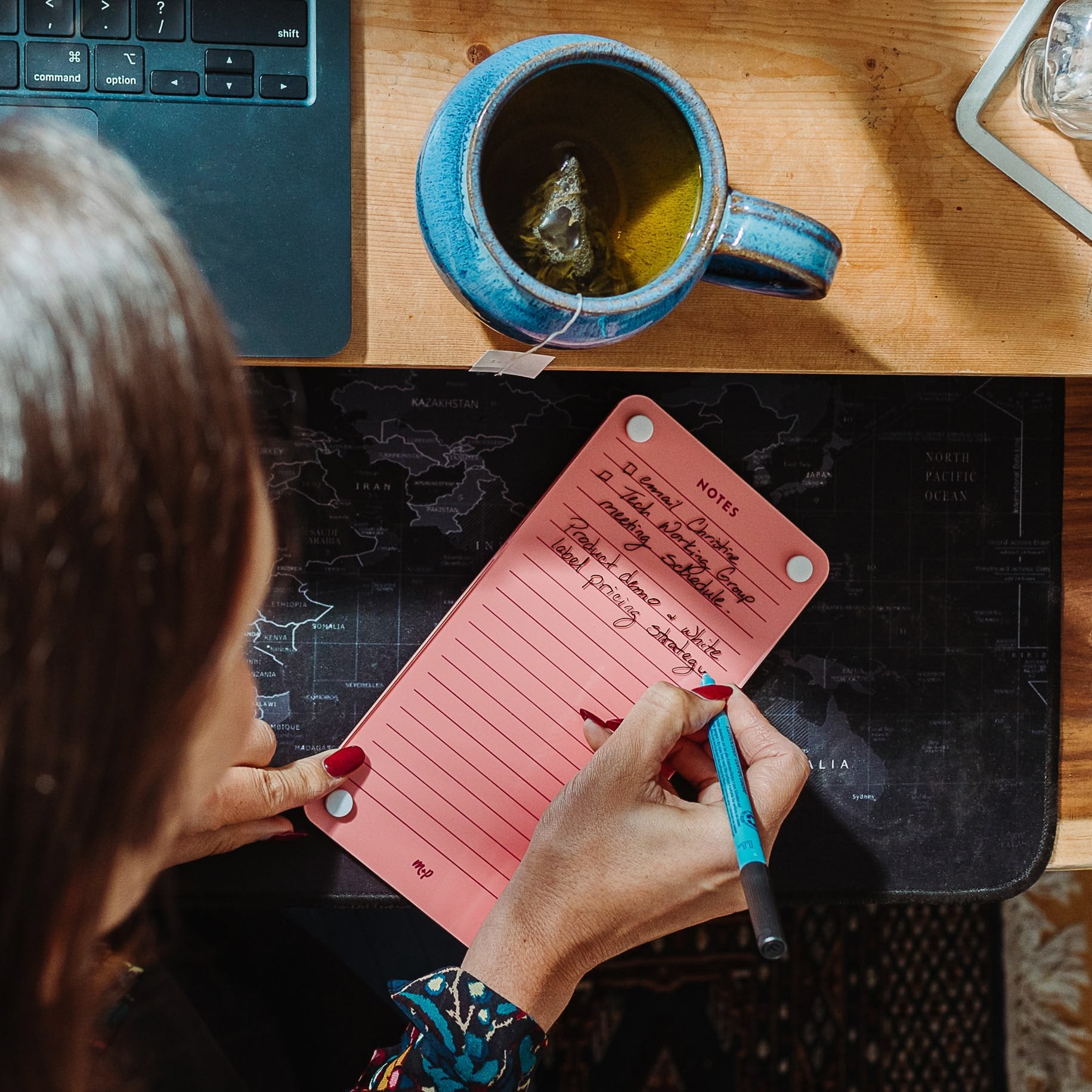 A person writes meeting notes on the Monkey and Pebbles Lined Notes 59 pink notepad at a desk with a laptop, blue mug of tea, and black map mouse pad. The top-down photo shows their hand holding a turquoise pen.