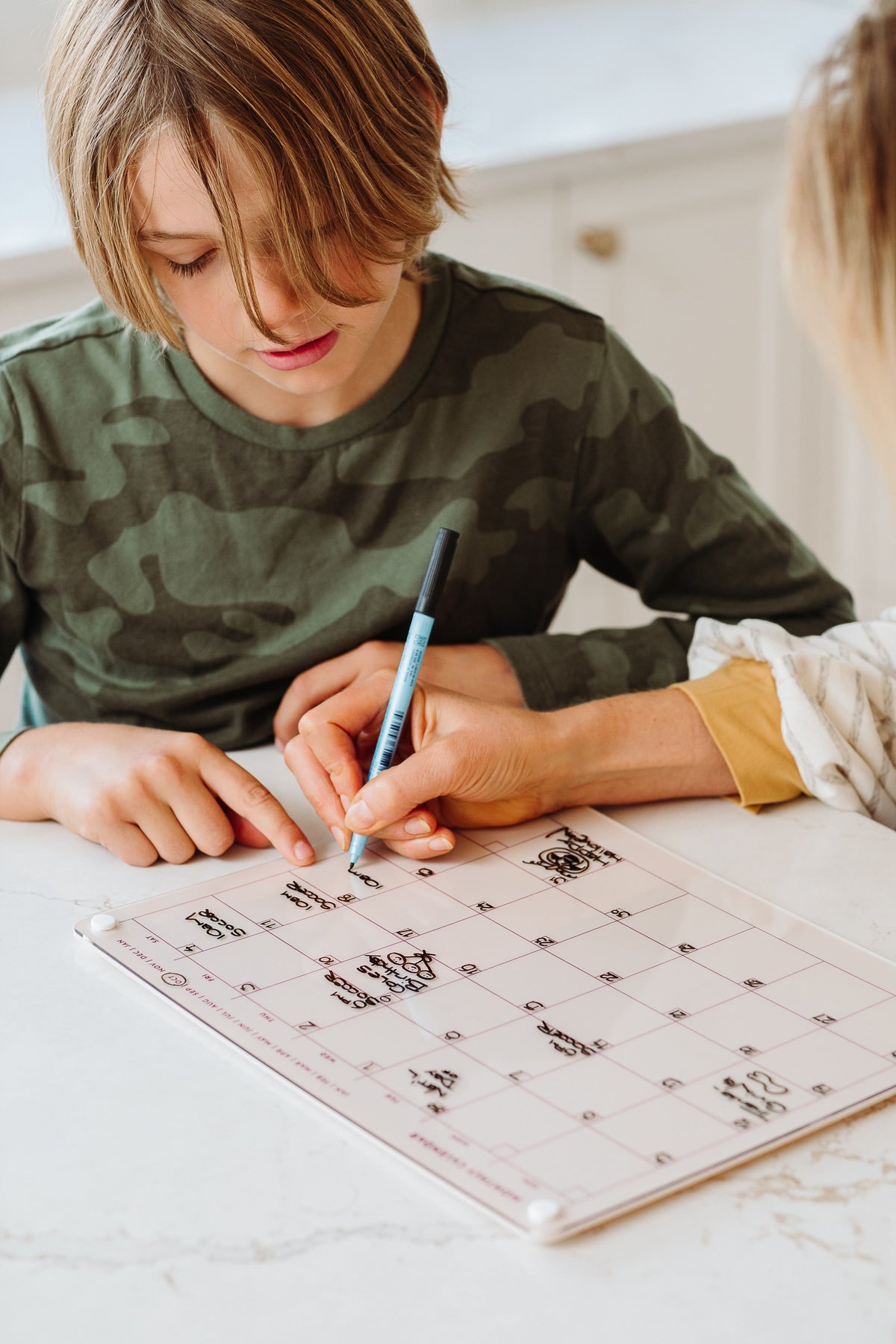 Boy and parent writing on a Linen magnetic dry erase monthly calendar on a kitchen counter