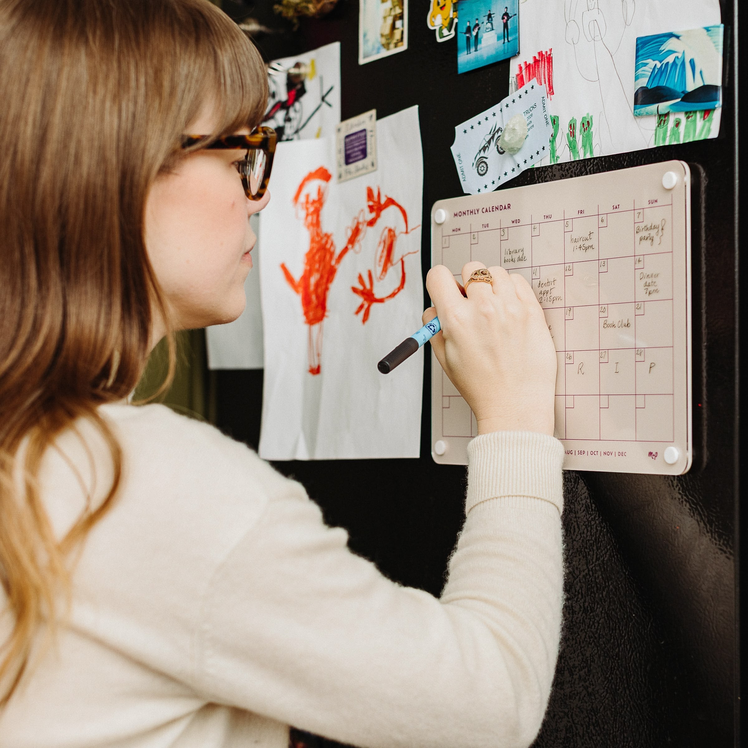 A woman writes on the Monkey and Pebbles Monthly Calendar 118, a compact planner attached to her refrigerator adorned with children’s drawings.