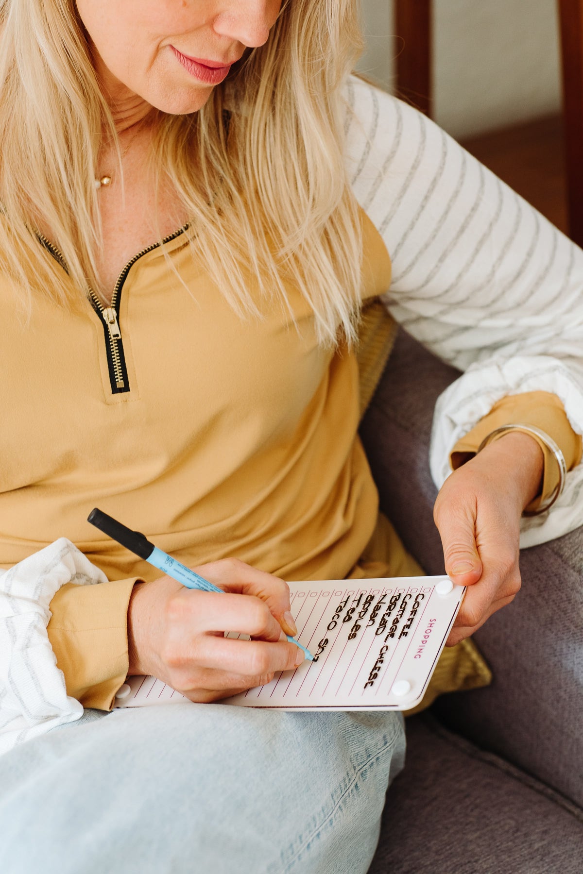 Woman writing her weekly grocery list on a Linen magnetic dry erase shopping list board while sitting on a sofa