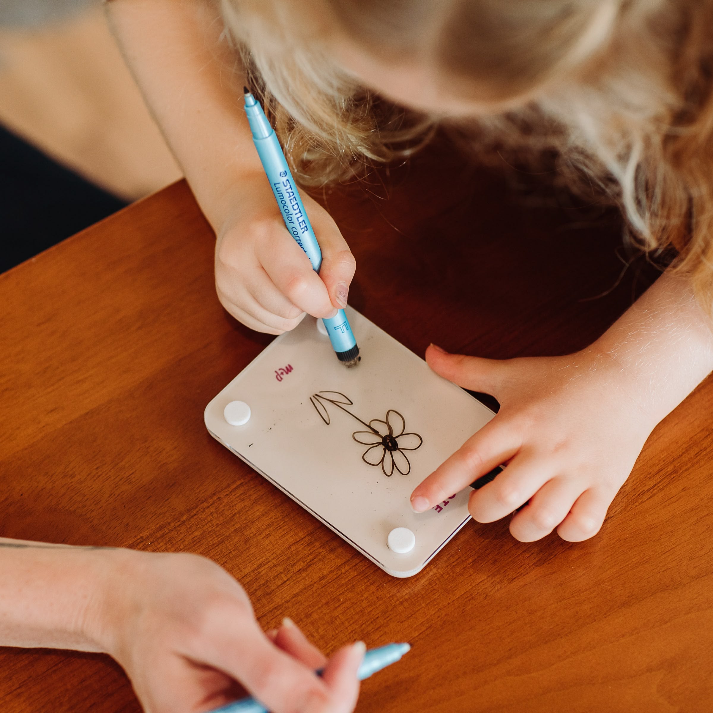 A child uses a blue marker to draw on the Monkey and Pebbles Little Note 35 drawing board atop a wooden table, while a blue pen appears nearby, making the scene resemble a modern sticky note.