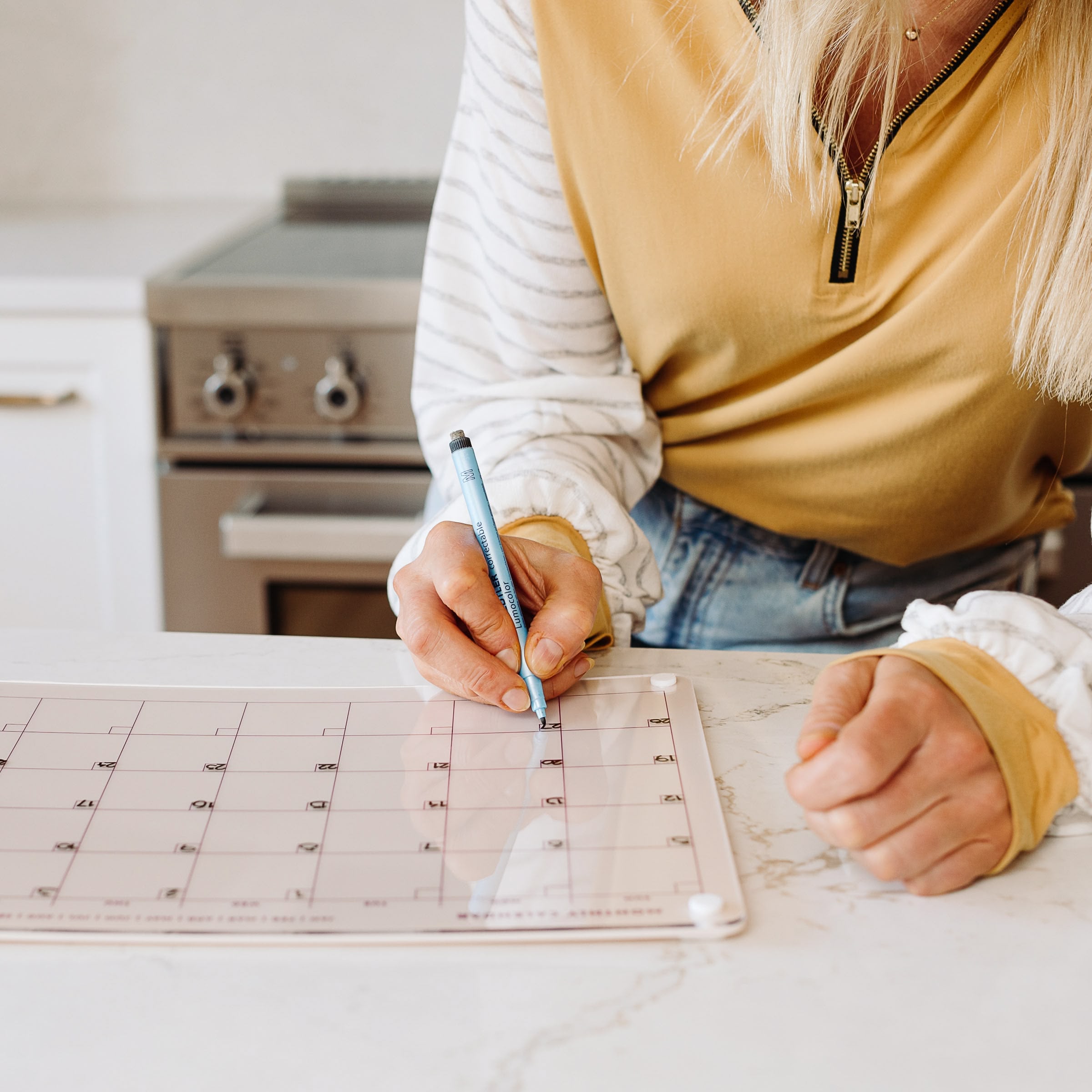 A person writes on the Monkey and Pebbles Monthly Calendar 1411 placed on the kitchen counter, with a stove in the background, while wearing a yellow and white long-sleeve shirt.