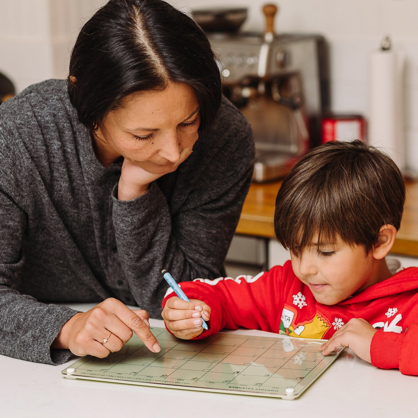 A woman and a boy in a red hoodie sit at a table, planning together with the Monkey and Pebbles Monthly Calendar 1411. The woman points to a date as the boy uses a blue marker to track their family plans.