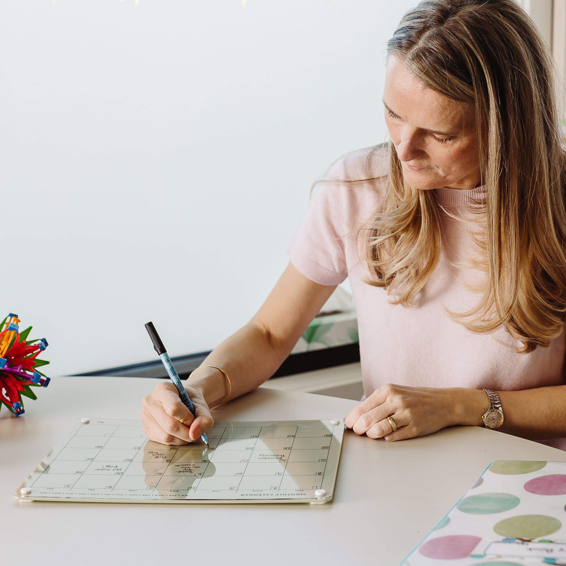 A woman with long blonde hair sits at a desk, writing on the Monkey and Pebbles Monthly Calendar 1411 with a black marker. The brightly lit desk is scattered with colorful items, as sunlight streams through the nearby window.