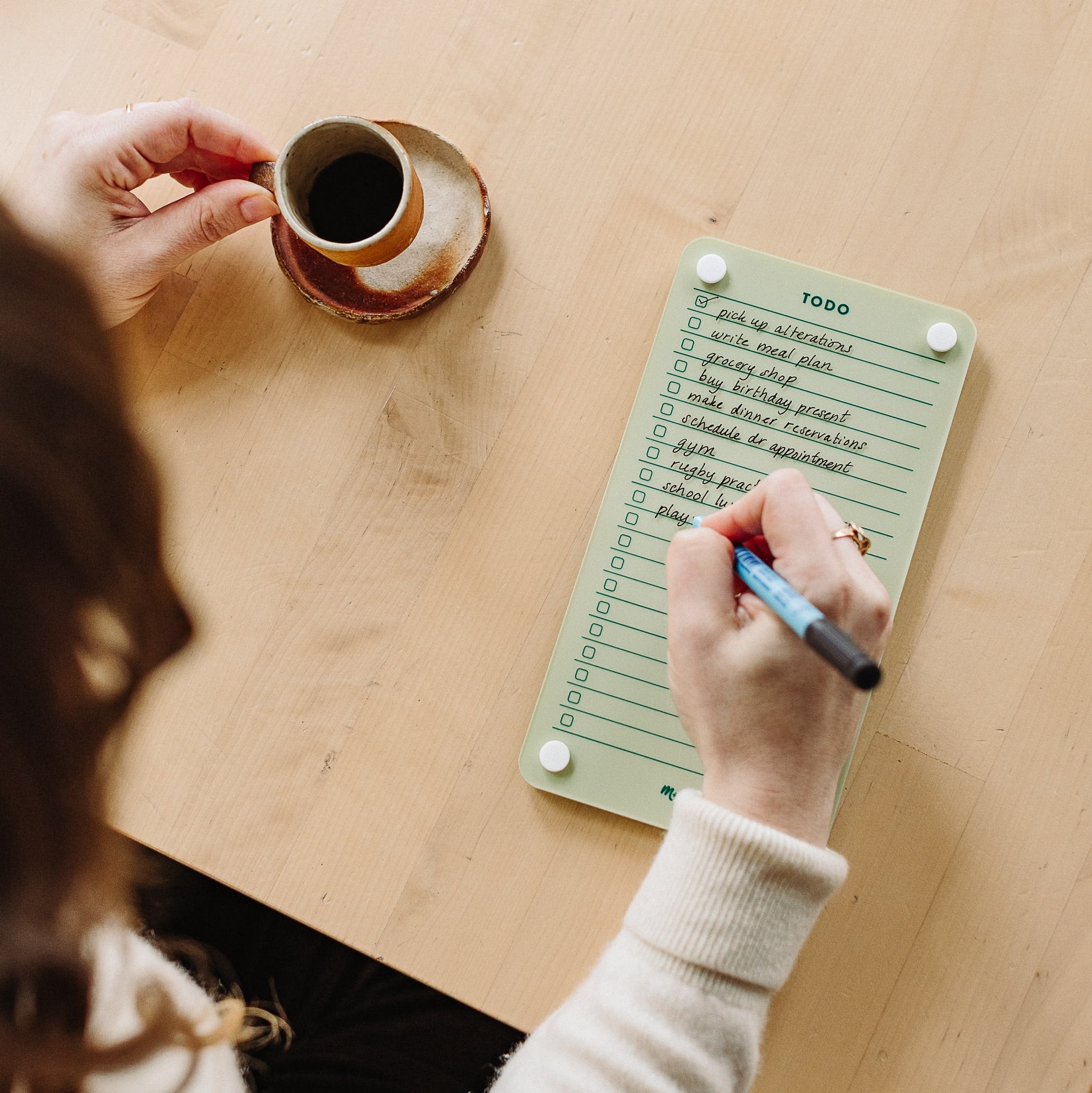 A person holds a cup of black coffee while writing on a Monkey and Pebbles Todo List 59 daily planner at a light wooden table. The green notepad has multiple tasks written and checked off.