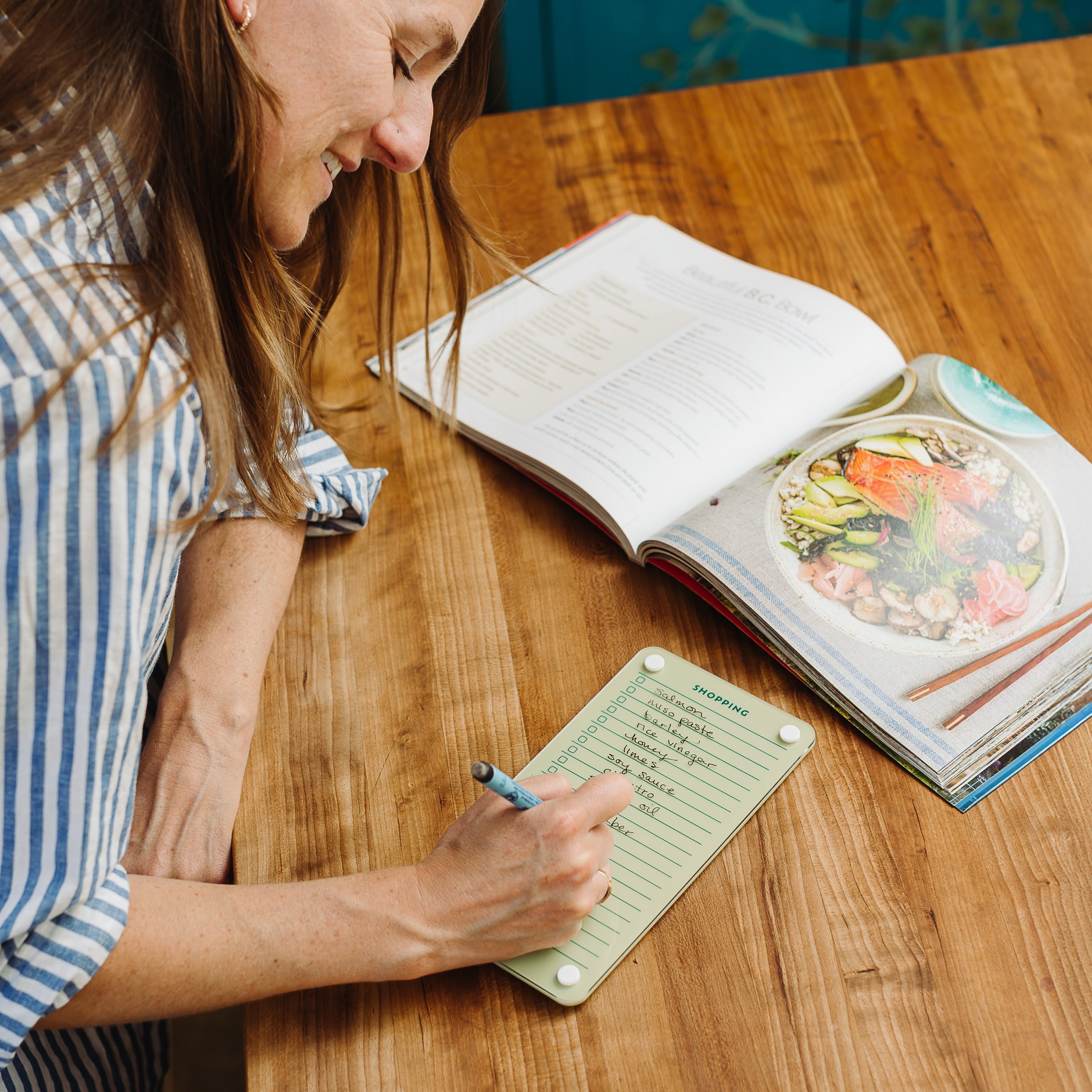 A woman smiles while writing a shopping list on the Monkey and Pebbles Everyday Home Bundle notepad at a wooden table, with an open cookbook beside her to help organize your home.