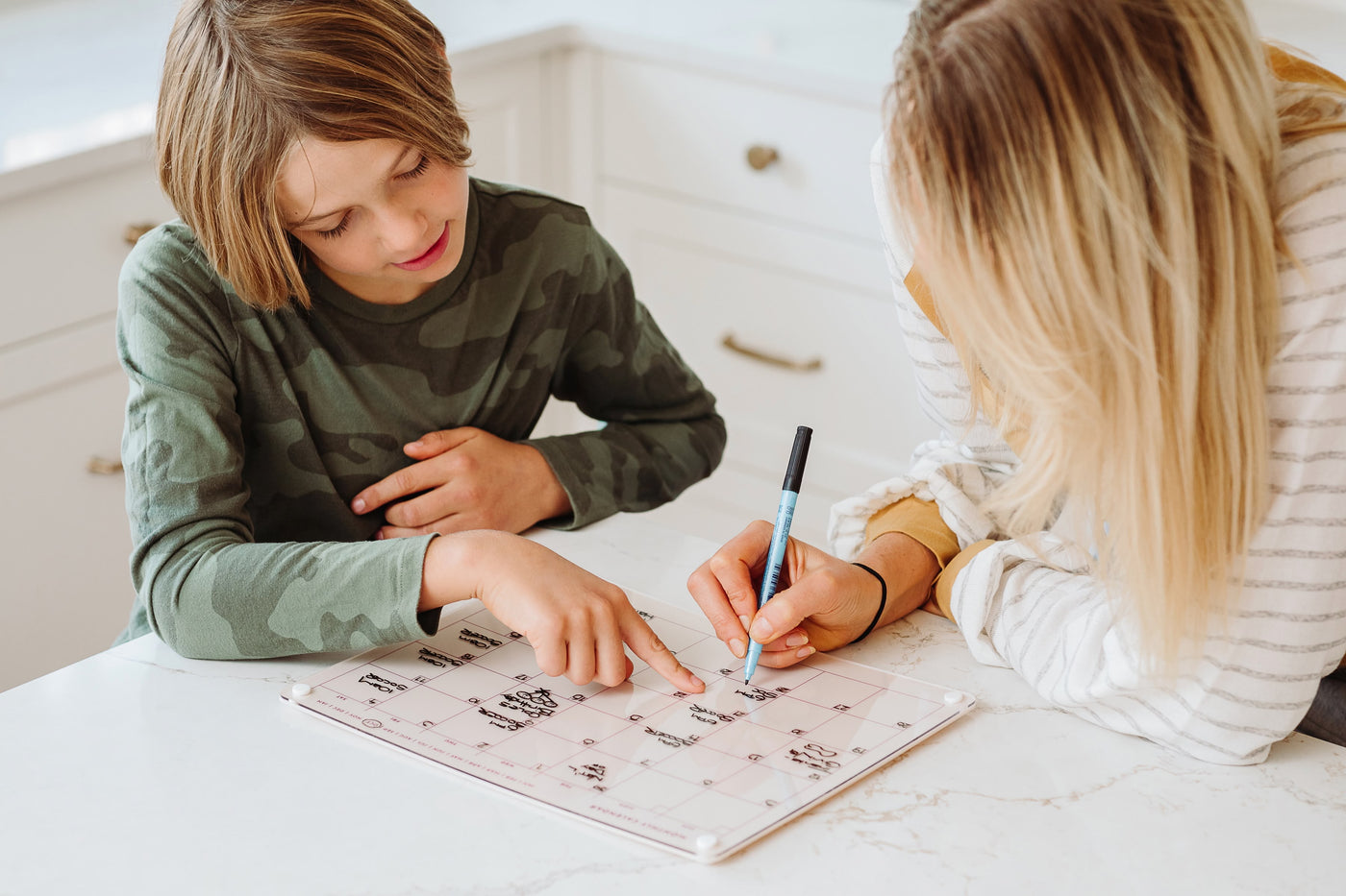 Mother and son using a Monkey and Pebbles magnetic monthly calendar to plan their schedule together at a modern kitchen counter