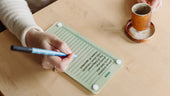 Flat lay of a hand writing on a Monkey and Pebbles sage green magnetic Todo List board next to a handmade ceramic coffee cup