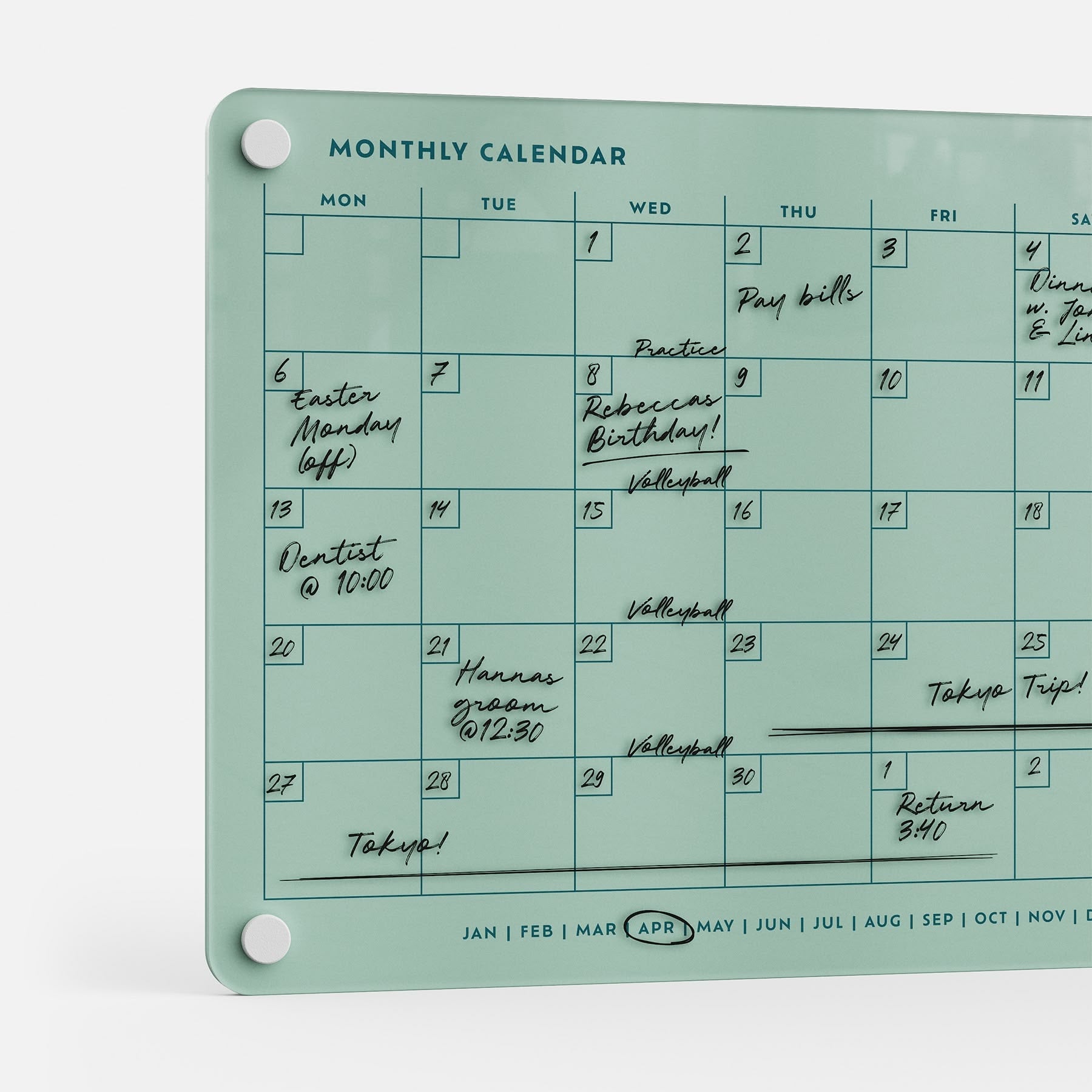 The Monthly Calendar 118, a compact glass dry-erase planner for April, is wall-mounted and filled with handwritten events like bills, dentist visits, volleyball, birthdays, and a Tokyo trip. White magnets secure each corner.