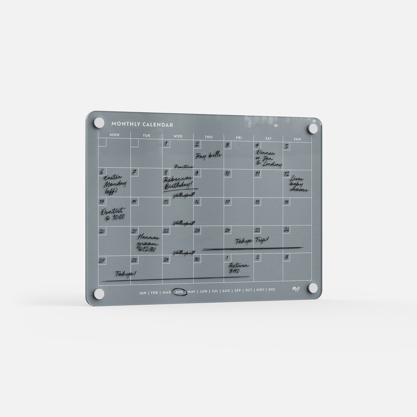 The Monthly Calendar 118 is a gray, compact wall planner with four corner holes and a minimalist design. Handwritten events and tasks in black marker fill each day, and month abbreviations are displayed along the bottom. Mounted on a white wall.