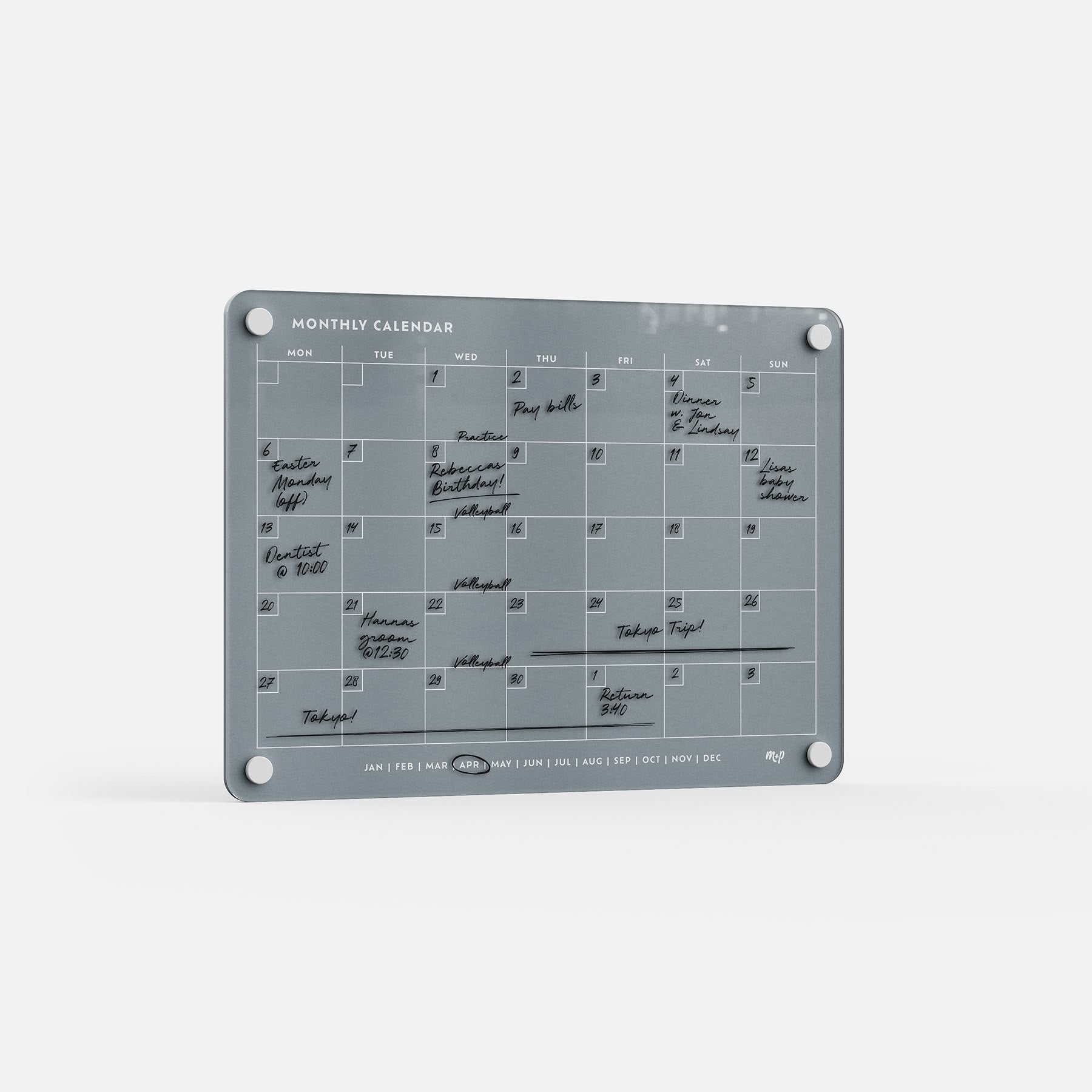 The Monthly Calendar 118 is a gray, compact wall planner with four corner holes and a minimalist design. Handwritten events and tasks in black marker fill each day, and month abbreviations are displayed along the bottom. Mounted on a white wall.