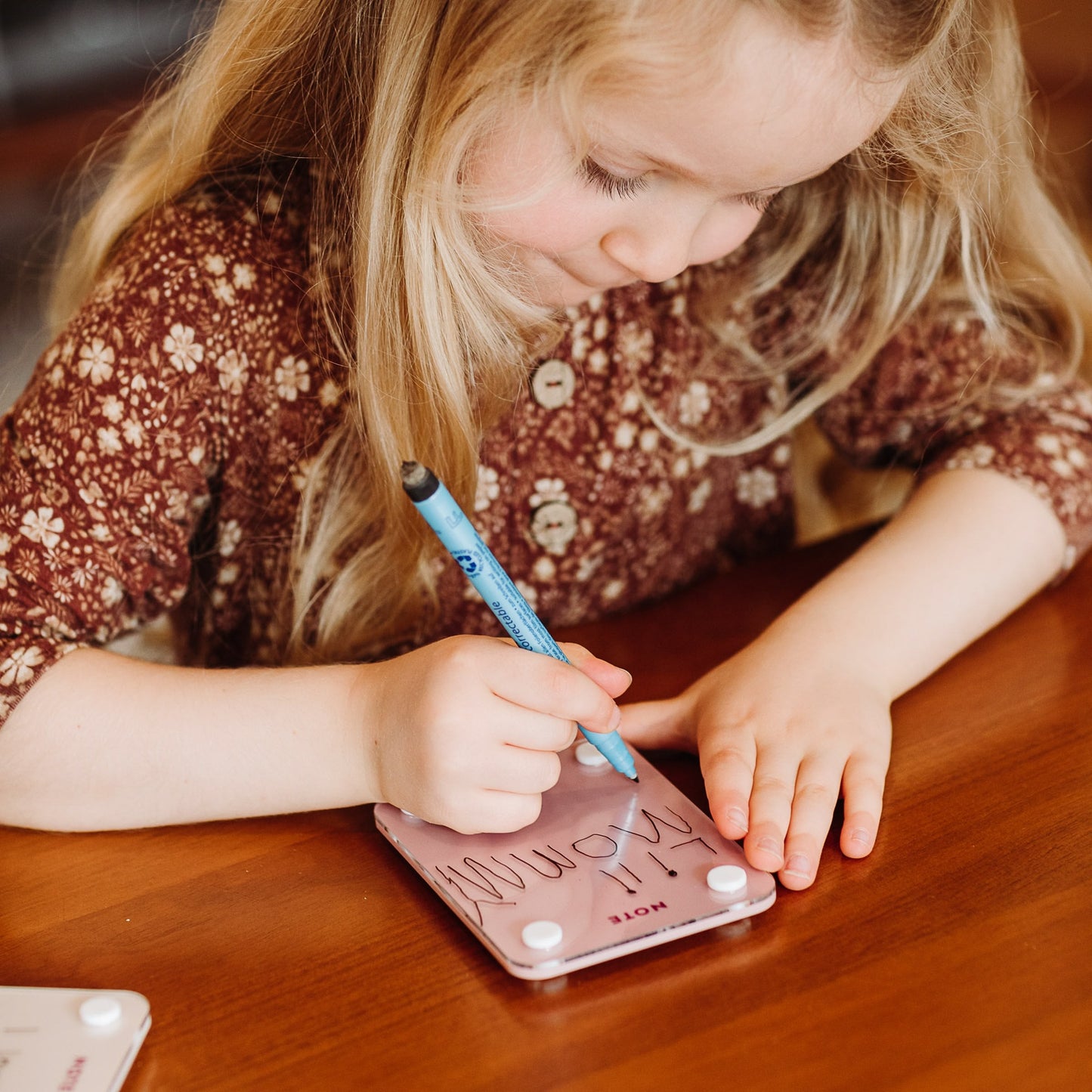 A young girl in a brown floral dress focuses as she writes a reminder with a blue marker on her Monkey and Pebbles Little Note 35 sticky note at a wooden table.