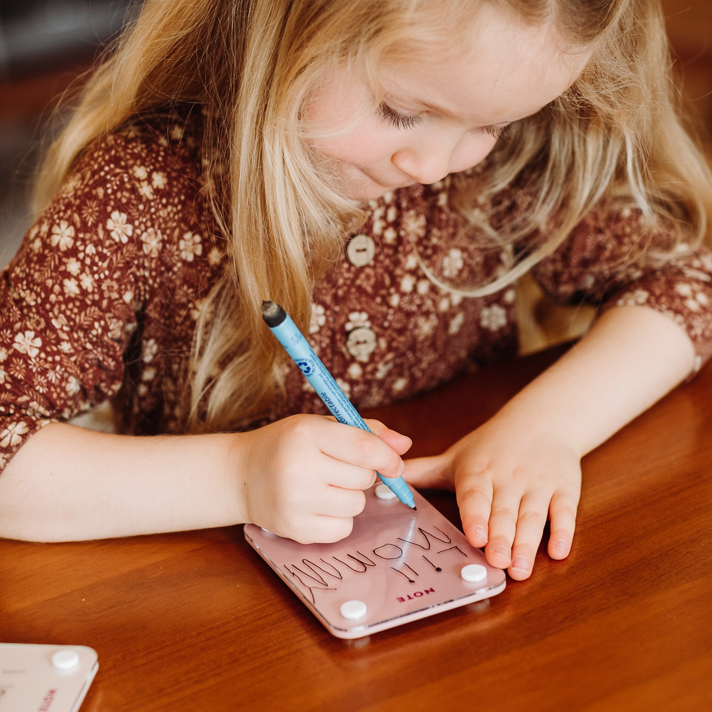 A young girl in a brown floral dress focuses as she writes a reminder with a blue marker on her Monkey and Pebbles Little Note 35 sticky note at a wooden table.
