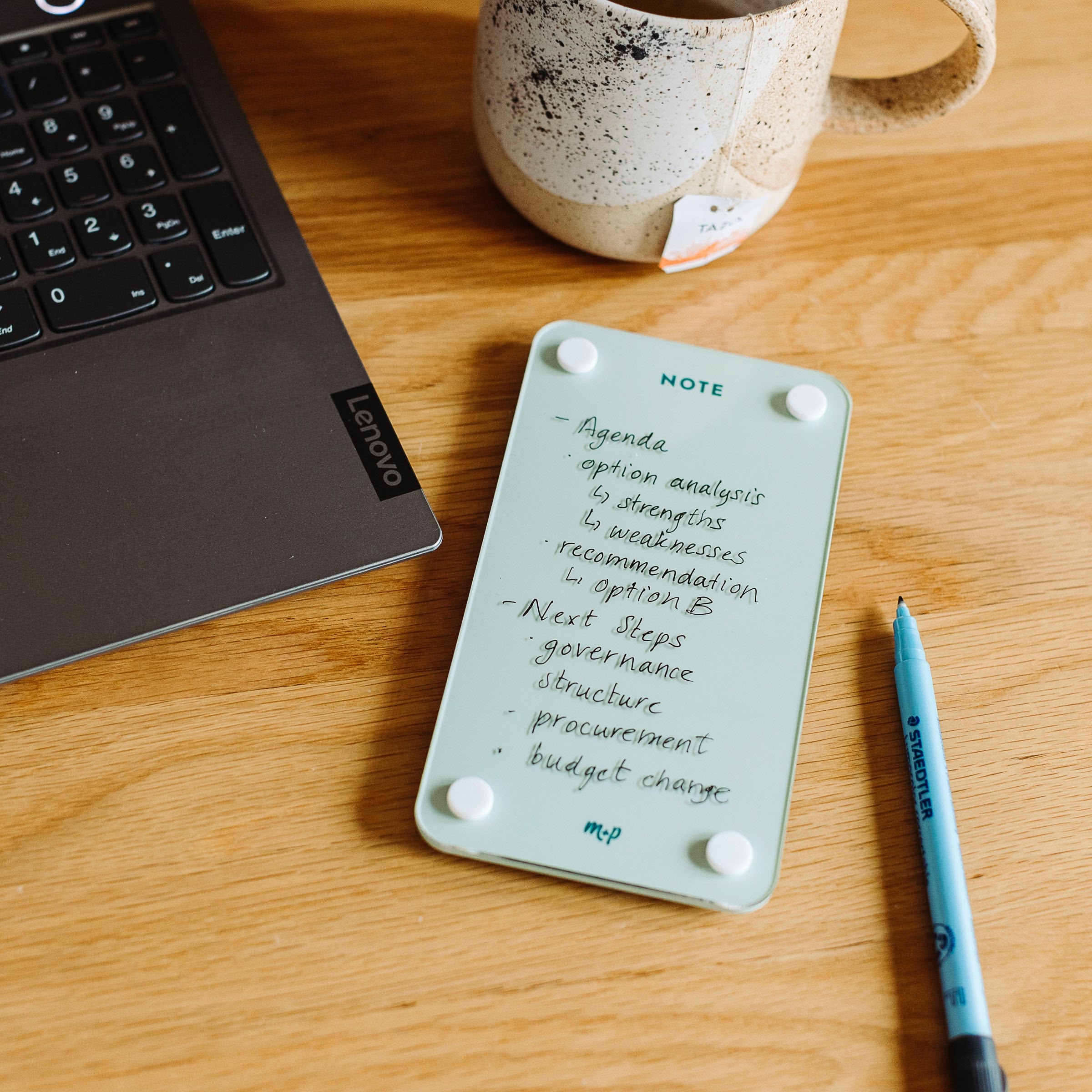A Monkey and Pebbles Tall Note 47 notepad with handwritten tasks sits on a wooden desk beside a Lenovo laptop, blue pen, and ceramic mug—ideal as a message board for agenda items like option analysis and budget changes.