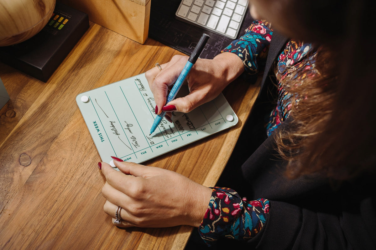 Woman writing on a Seafoam magnetic dry erase weekly planner board at a wooden desk beside a keyboard