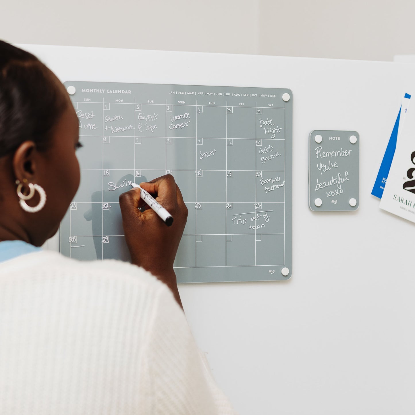 Someone writes on the gray magnetic monthly calendar from the Everyday Home Bundle by Monkey and Pebbles. Nearby, a matching notes board displays "Remember you're beautiful," while clipped papers add to the organized setup.