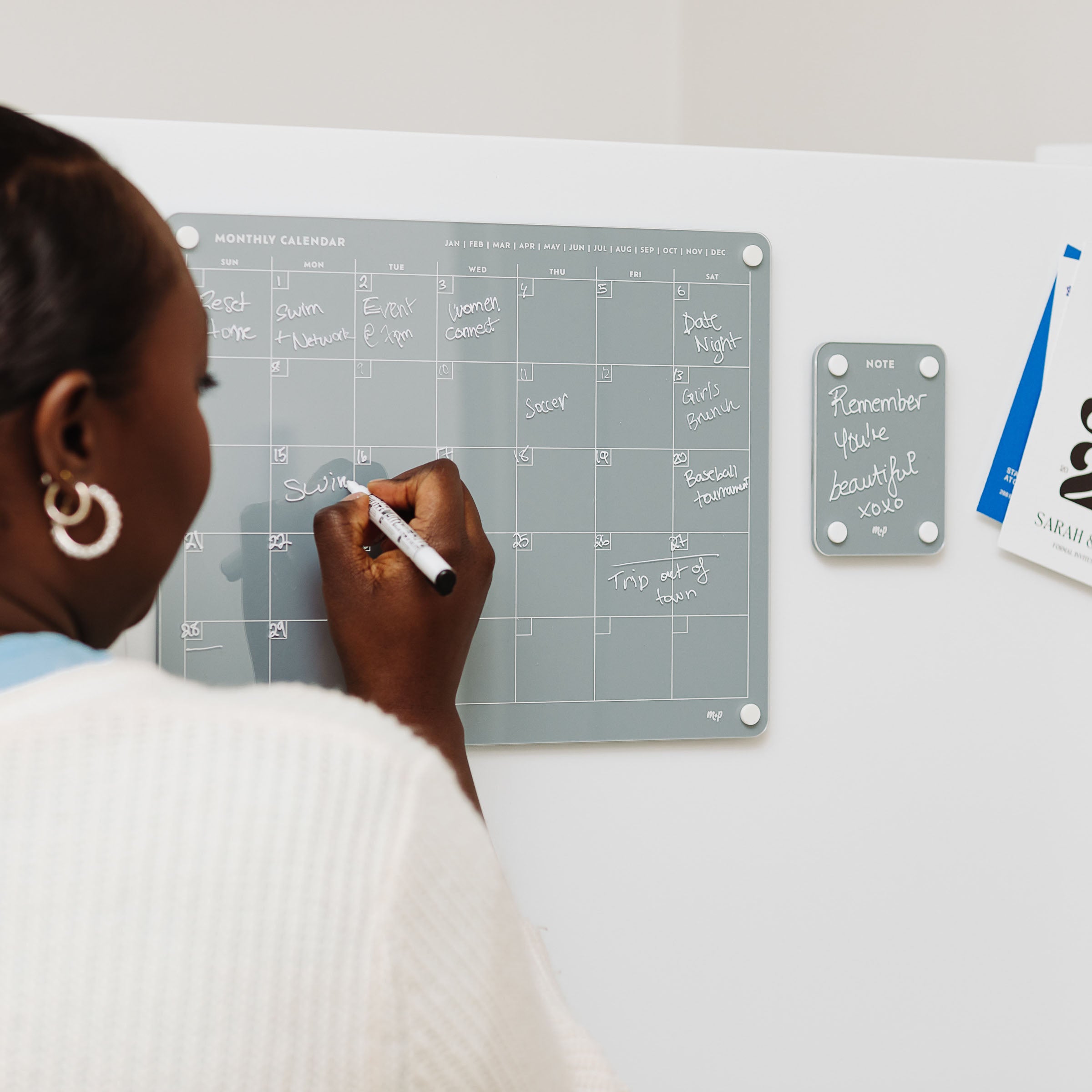 Someone writes on the gray magnetic monthly calendar from the Everyday Home Bundle by Monkey and Pebbles. Nearby, a matching notes board displays "Remember you're beautiful," while clipped papers add to the organized setup.