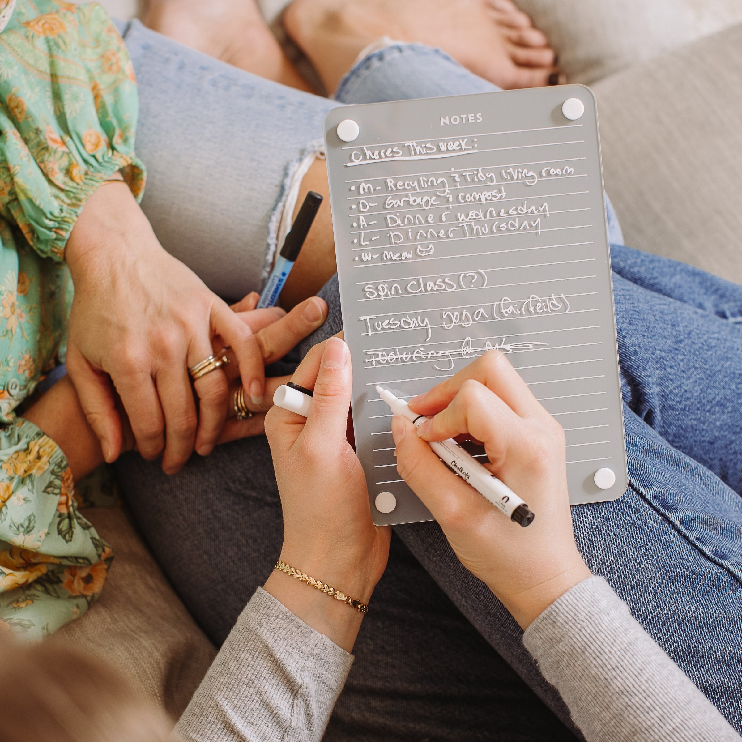 Two people sit together, using a gray Monkey and Pebbles Lined Notes 69 reusable planning board and stylus to jot weekly tasks—recycling, garbage, dance, dinner, spa class, yoga. Only their hands and laps are shown.