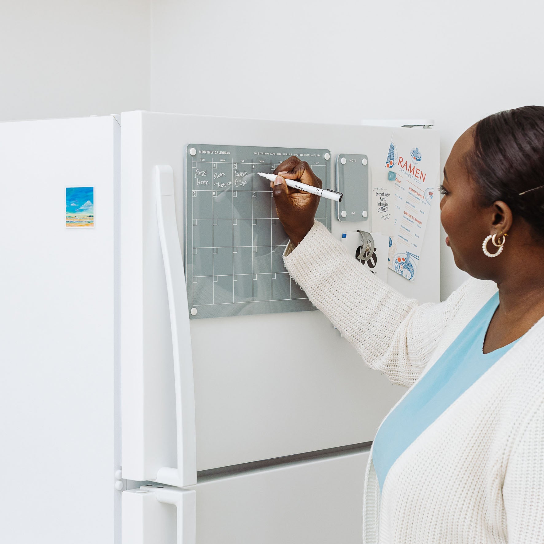 A woman wearing a sweater and gold hoop earrings organizes her schedule with the Monkey and Pebbles Monthly Calendar 1411, attached to her refrigerator alongside a postcard and a paper titled RAMEN.