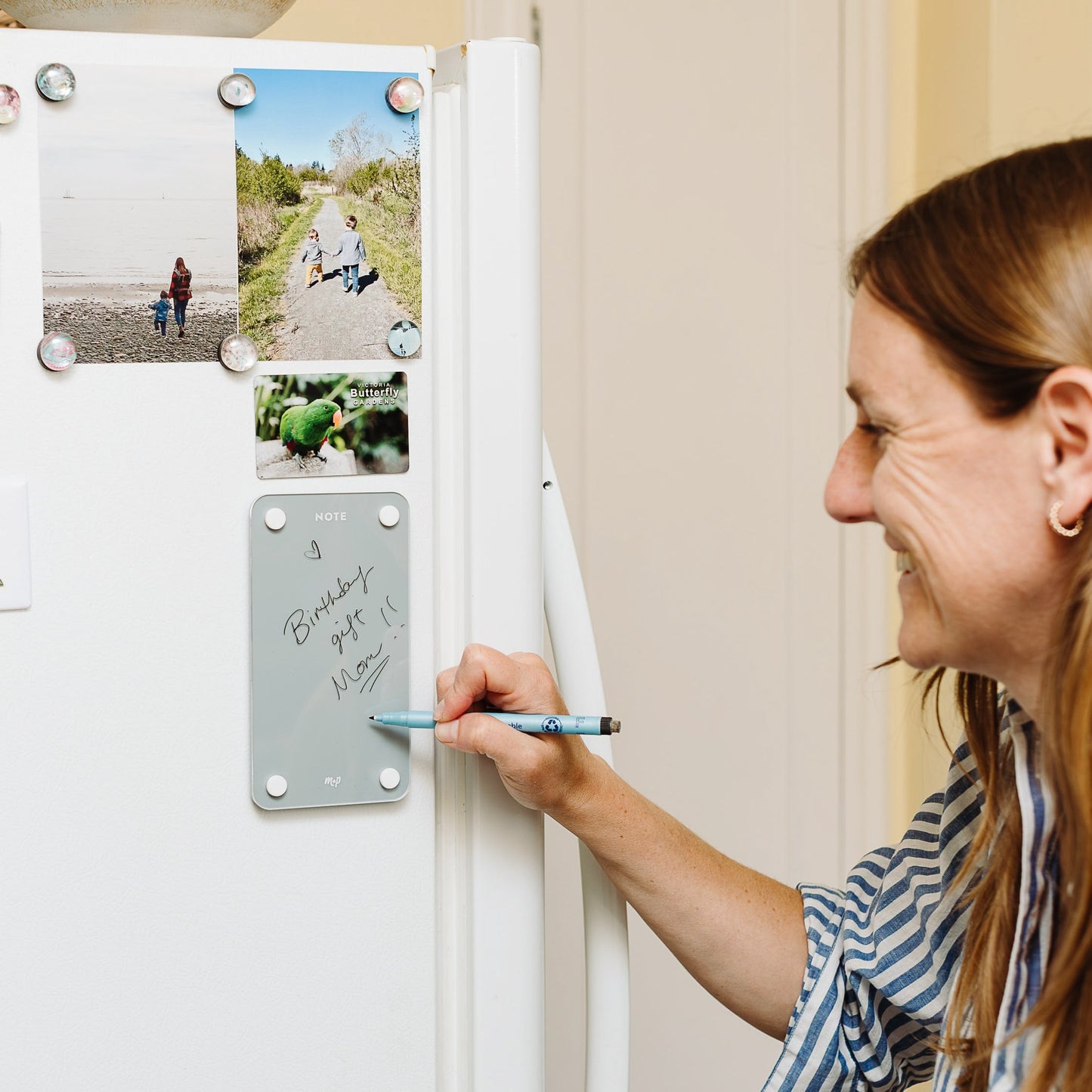 A woman smiles as she writes "Birthday gift - must!!" on the Monkey and Pebbles Tall Note 47 magnetic dry-erase board attached to her fridge, using it for quick lists and reminders alongside photos.