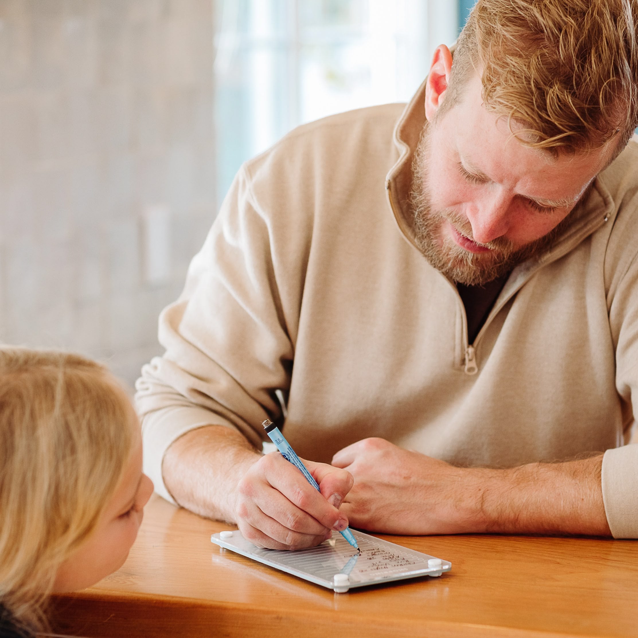 A man with a beard writes on a Monkey and Pebbles Shopping List 59 notepad while a young blonde girl watches from across a wooden table.