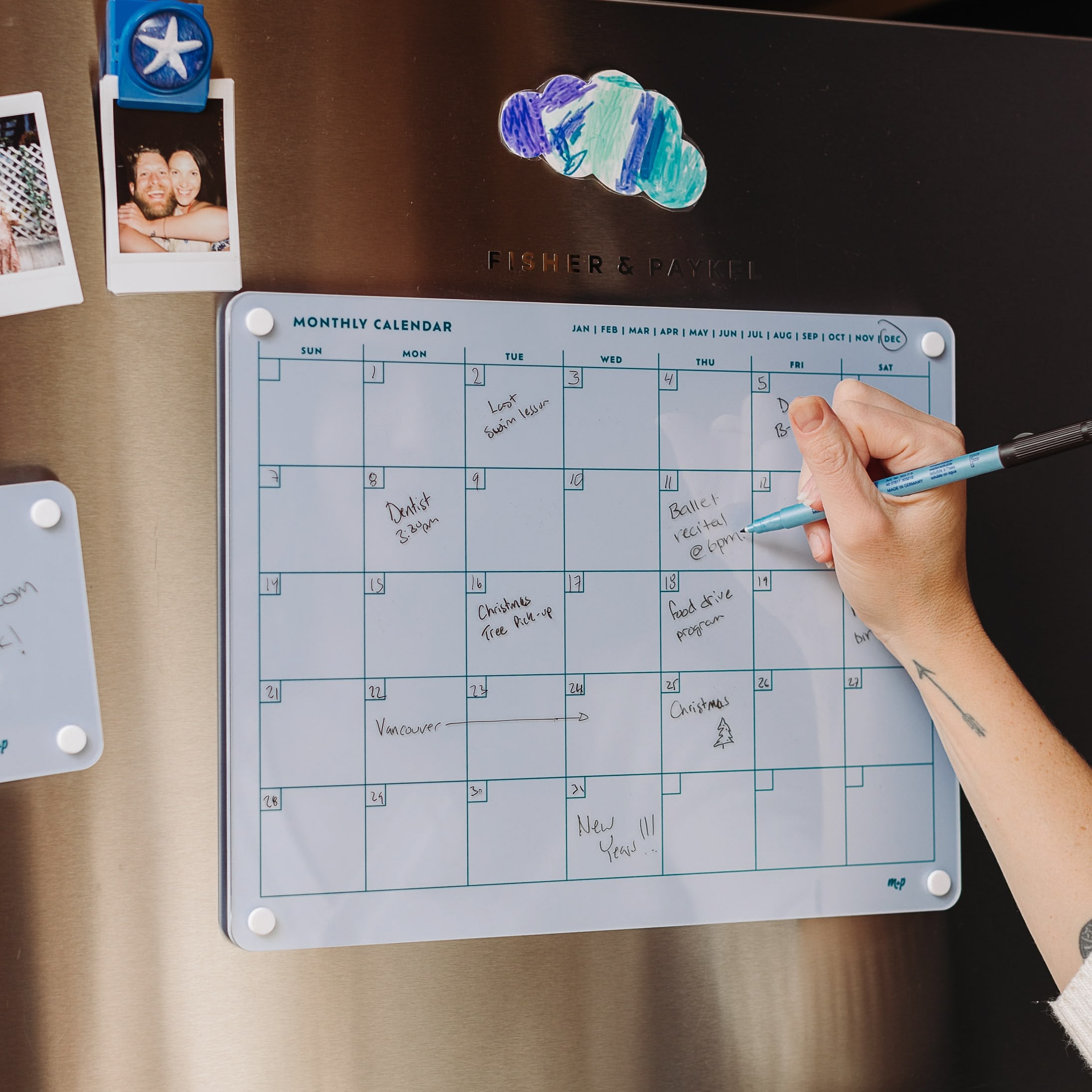 A person writes on the Monkey and Pebbles Monthly Calendar 1411, a blue dry-erase planner attached to a stainless steel fridge, surrounded by family photos and featuring handwritten notes for organizing events.