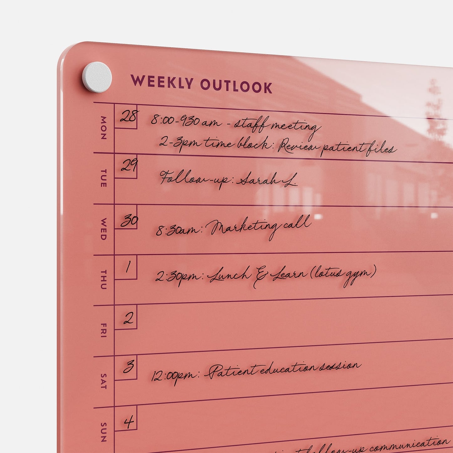 The Weekly Outlook 811 is a pink dry-erase weekly planner board attached to the wall by a round magnet, listing days of the week on the left. It helps organize handwritten tasks like staff meeting, calls, lunch & learn, and patient education sessions.