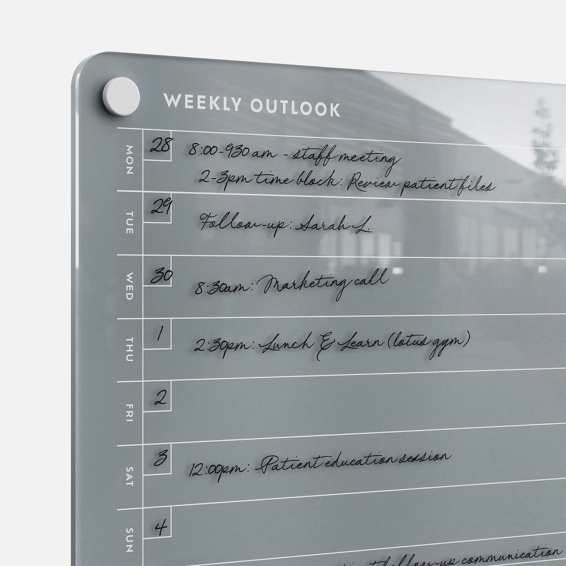 A close-up of the Weekly Outlook 811 glass dry-erase board, a stylish weekly planner and productivity organizer, displays handwritten events from Monday to Saturday in black ink and features magnetic fasteners in the corner.